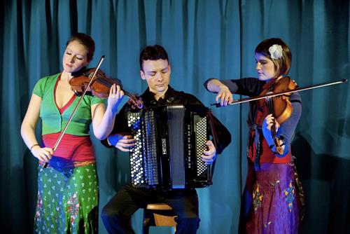 three musicians infront of a large curtain, two girls playing violins and youn man sat playing the accordion